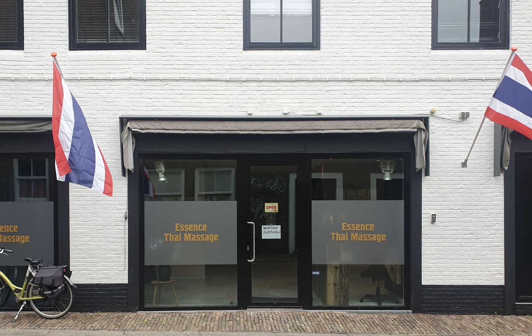 Witte gevel aan de Sint Janstraat met de ramen van Essence Thai Massage en twee Thaise vlaggen