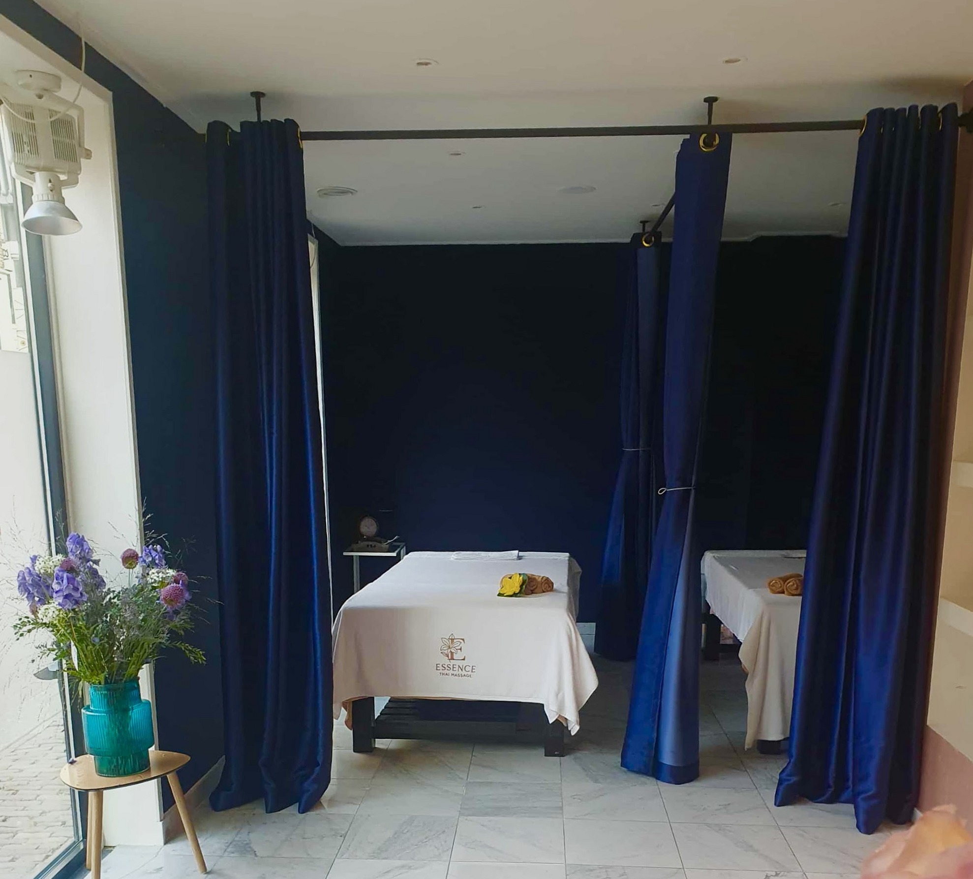 Behandelruimte met twee massagetafels achter donkerblauwe gordijnen en een vaas met blauwe bloemen